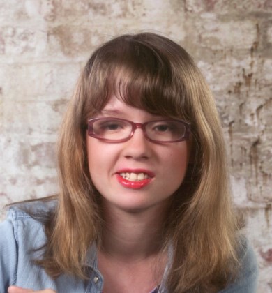 head and shoulders photo of Sarah Cornett. Sara is a young adult white woman with blonde hair just beyond her shoulders. She is wearing glasses and is posed in front of a white washed brick wall