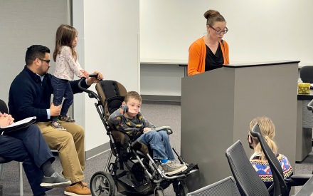 an advocate mom speaks from behid a lectern, while her husband sits behind her, holding a stroller with their son inside