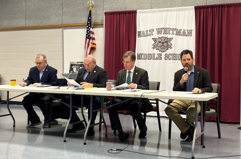Four male elected legislators sit behind a long folding table in a school gymnasium