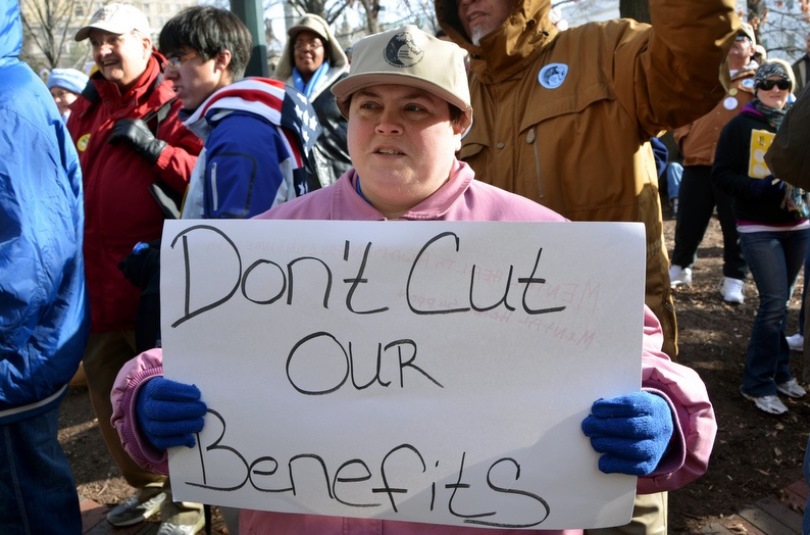 Self advocate outdoors in a coat, hat and mittens holding a sign that reads Don't cut our benefits