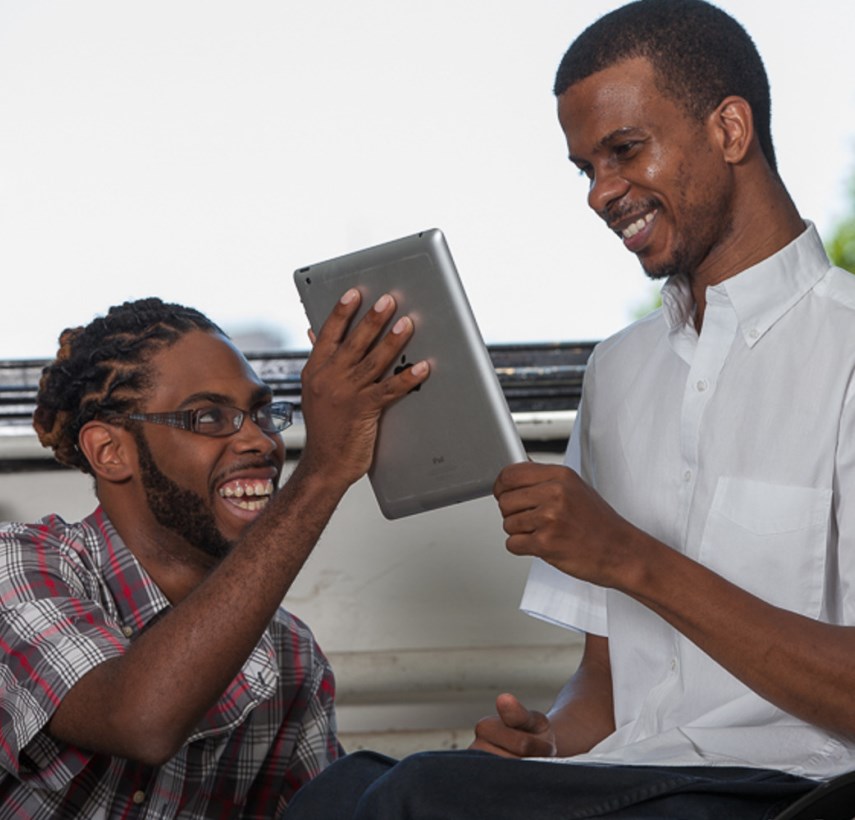 Two Black men smiling; one is showing the other an iPad.