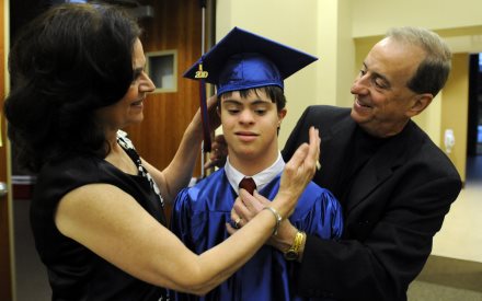 a young man with DD stands in a graduation cap and gown, while his mom and dad look on