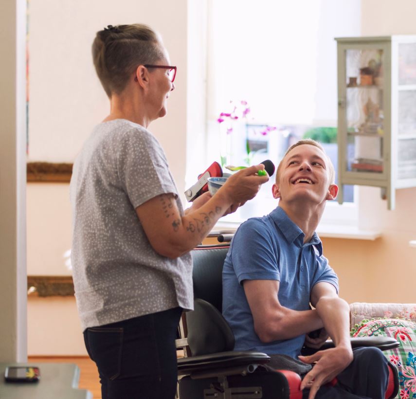 A young white man in a wheelchair laughs as he interacts with an older white woman serving as his support coordinator