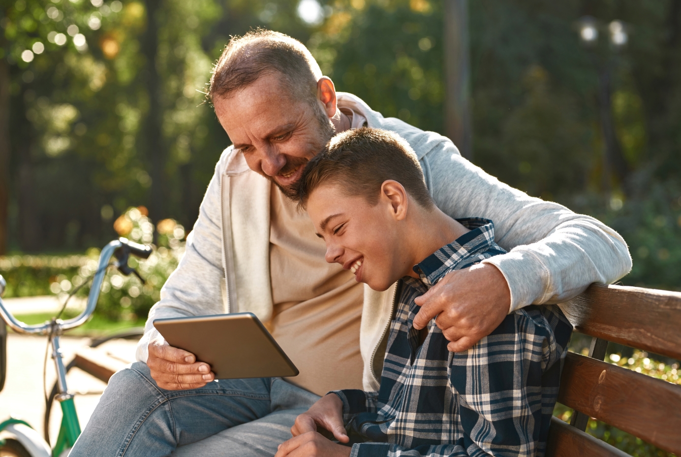 a man and his son with a developmental disability sit on a park bench outdoors. The father holds a tablet in his right hand, his left arm is around the boys's shoulders.