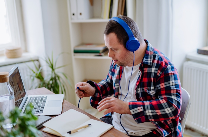 a man with a developmental disability, perhaps in his late 20s or 30s, sits at a table, wearing headphones, and about to plug the headphones into a laptop computer on the table.