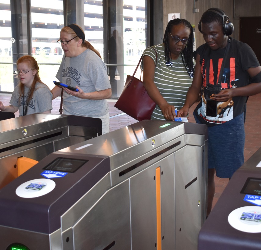 a young adult female with IDD and her mother pass through a MetroRail faregate. To the righ, a young adult Black male with IDD and his other pass through another faregate.