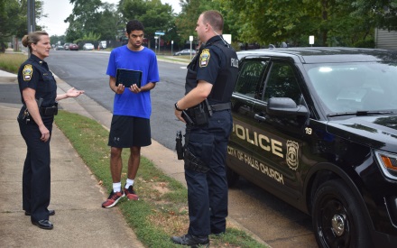 outdoors on a city street, a young man with IDD speaks with two police officers, standing beside their squad car.