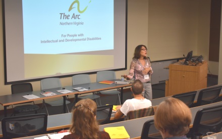 A presenter stands in front of a classroom. Behind the presenter a powerpoint slide showing The Arc of Northern Virginia's logo is on the screen on the wall. The heads of three attendees are seen in the foreground of the photo