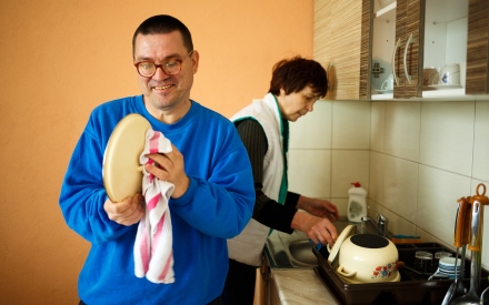 an adult male wearing a blue shirt dries a plate with a dishtowel. Behind him his mother washes dishes in the sink.