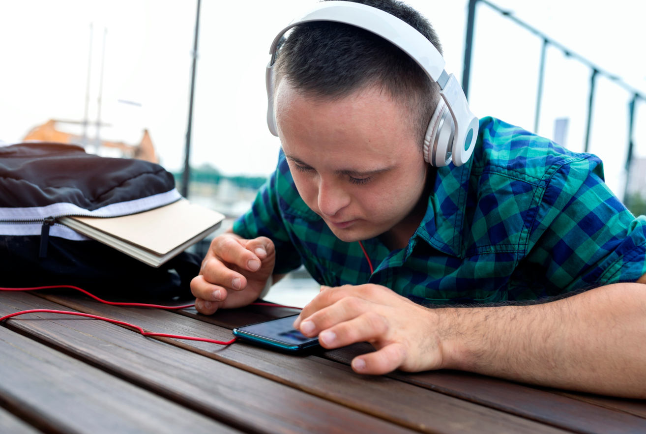 a young man in a blue striped shirt and wearing headphones uses a mobile device while sitting at a table.