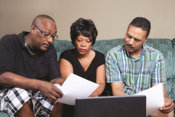 Three members of an African-American family, father, mother, and adult son, sitting on a couch reviewing documents in front of an open laptop on the coffeetable