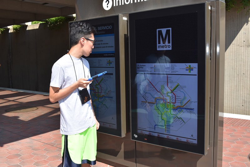 A young Asian man holding an iPad stands in front of a map of the Metro on an electronic display at a Metro rail station.