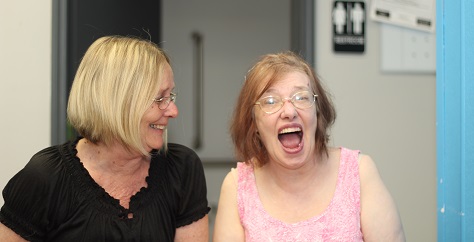 blonde woman sits next to and looks at a redheaded woman with a wide open-mouthed smile.