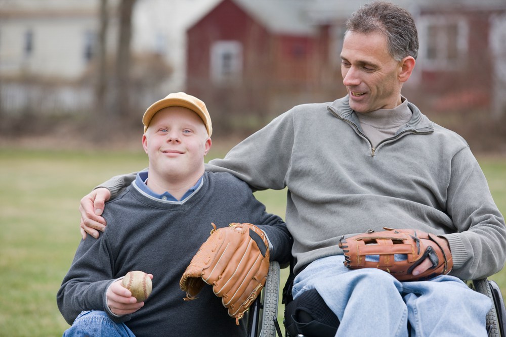 Dad in a wheelchair with a baseball mit, puts his arm around his son who has Down syndrome, and is also in a wheelchair with a baseball mit