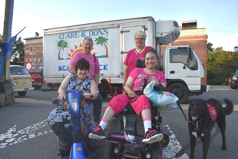two women with IDD in motorized wheelchairs, with a large black guide dog beside them, and two female friends standing behind the wheelchairs