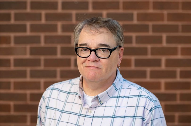 Head and shoulders photo of Rob Hudson, wearing glasses and a plaid dress shirt, posed in front of a red brick background.
