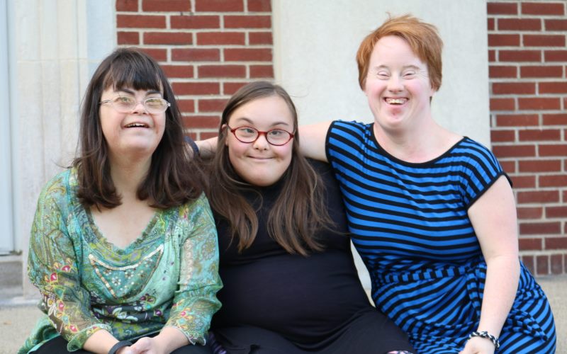 Three young adult women with Down syndrome, dressed in business casual blouses, sit with their arms around eah other on a bench in front of a red brick wall.