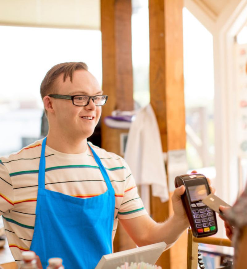 Young man with IDD wears a blue apron while working behind the counter of a coffee shop, holding a credit card machine for a customer.