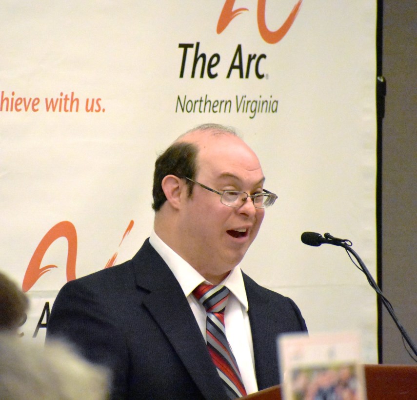 Self-advocate David Egan, wearing a suit coat and tie, speaks at a lecturn at The Arc of Northern Virginia benefit breakfast.