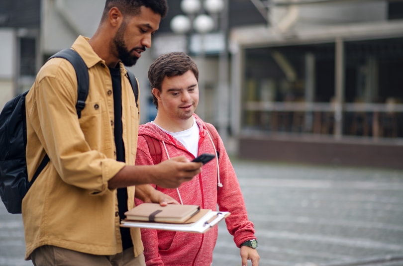 a young man with a develomental disability walks down the street with his mentor, a slighly older young man holding a smart phone.