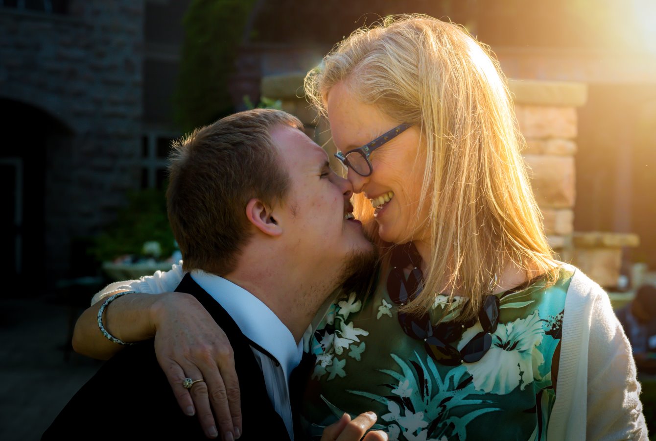 A mother and son wearing dress clothes stand looking at each other nose-to-nose, with sunlight illuminating them from the upper right corner.