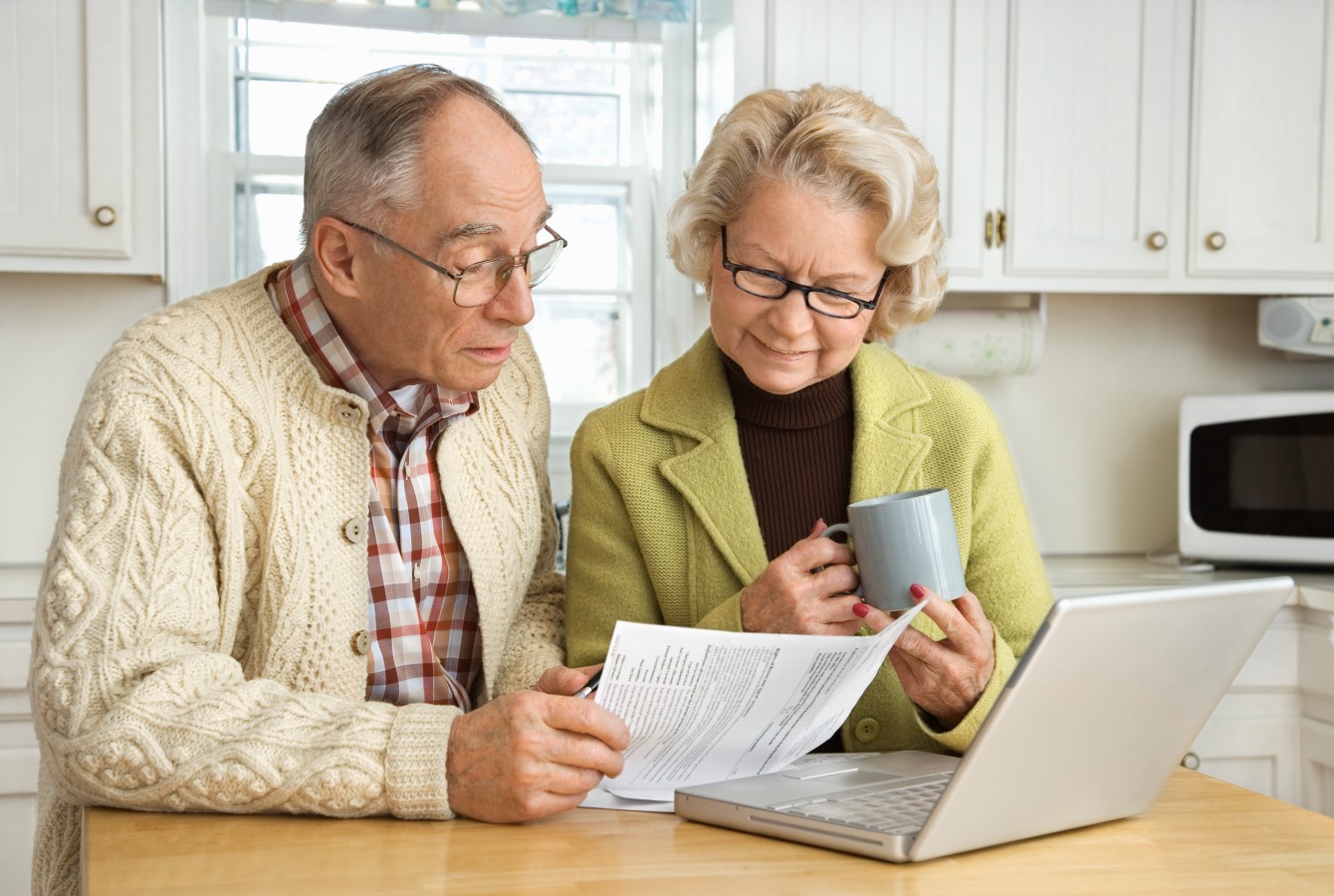 And older white couple look over a document in front of a laptop computer ona kitchen counter.
