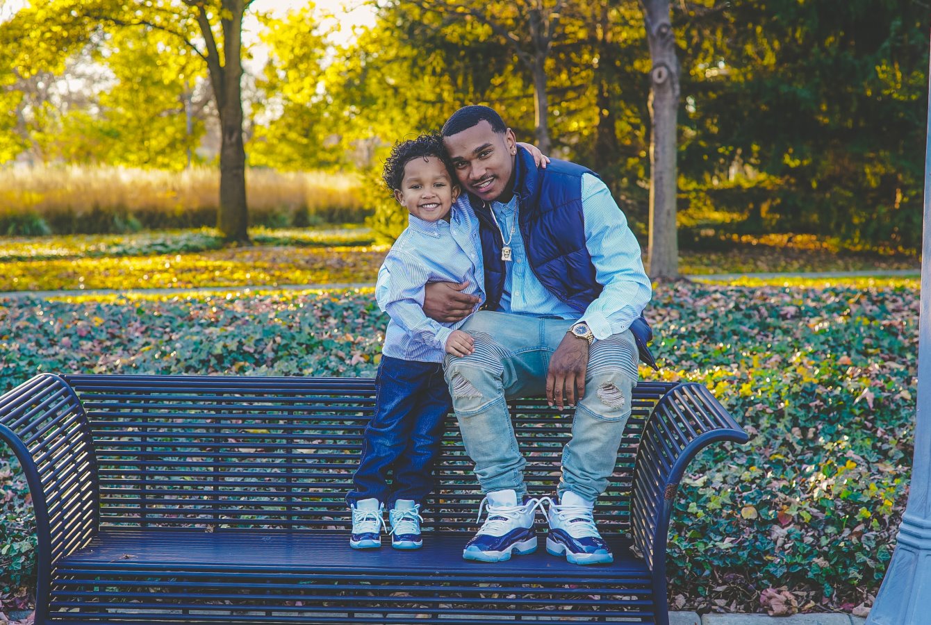A black father and his young son sit on the back of a park bench in front of trees with the leaves falling.