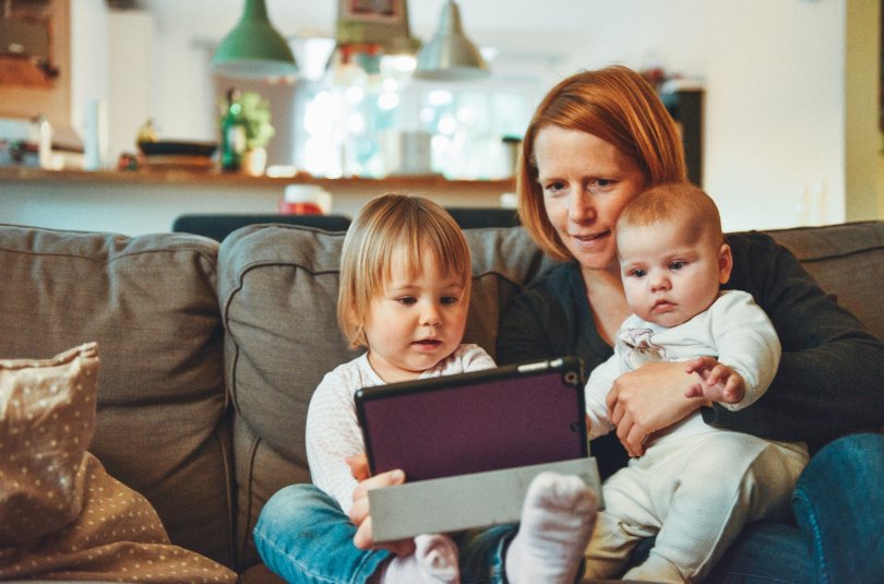 A young mom sits on the couch with a toddler and a baby, looking at a laptop screen