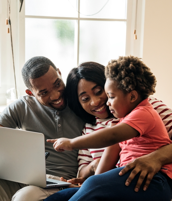 A young african american couple sits on the couch with their toddler son on mom's lap, pointing to a laptop screen that sits on dad's lap