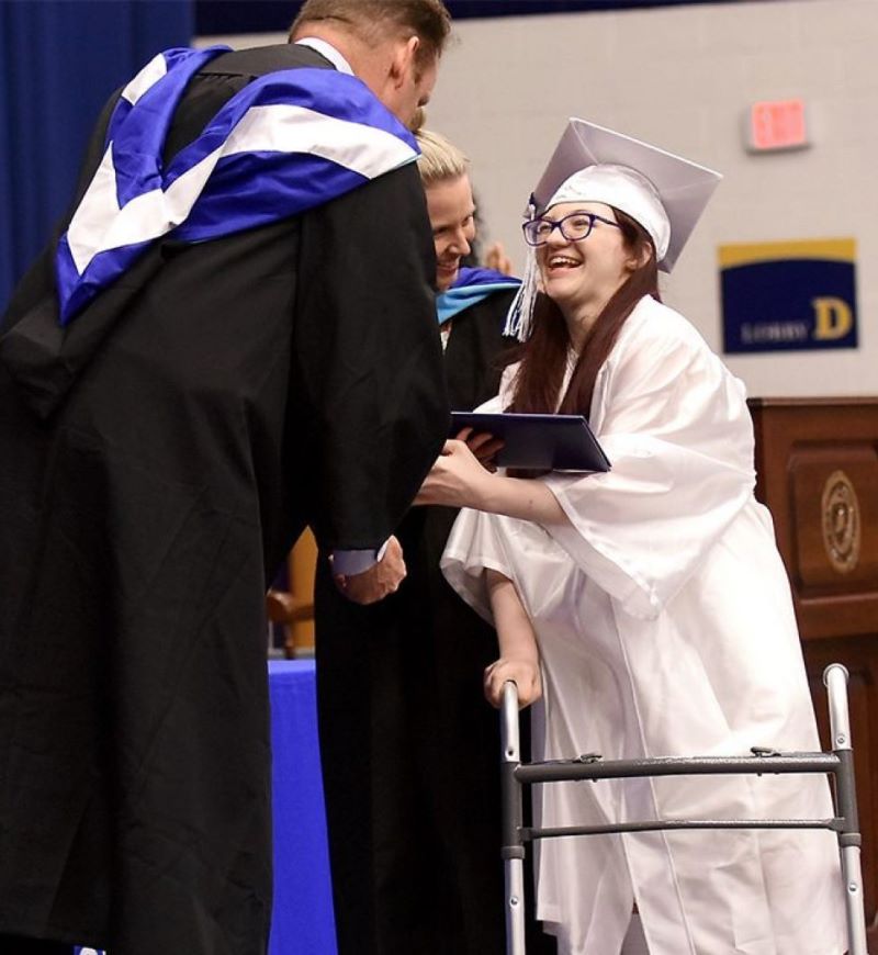 high school girl with disabilities, wearing a white cap and gown, stands with her walker, accepting her diploma from the superintendent, while her teacher looks on