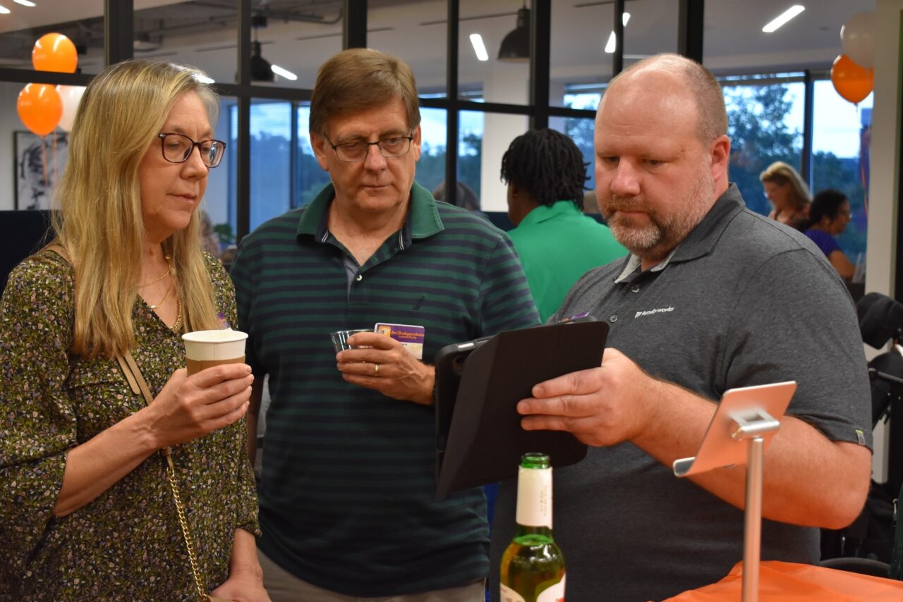 Photo shows a white woman with blonde hair wearing a green blouse, and a white man with glasses and a polo shirt looking at an iPad screen being held by another white man wearing a gray polo shirt