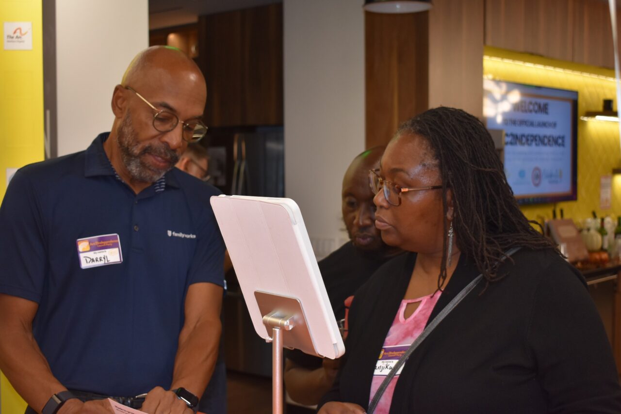 photo shows a black man wearing a blue polo shirt and a black woman wearing a black sweater looking at the screen of an iPad