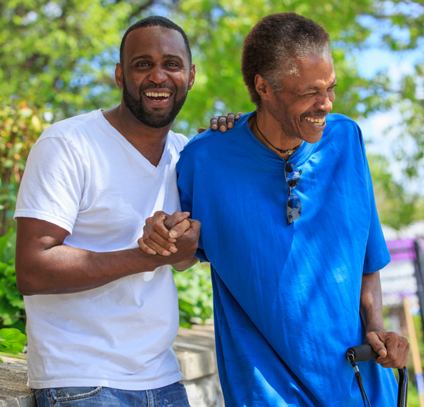 a young black man in a white t shirt steadies an older black man wearing a blue shirt and holding a cane