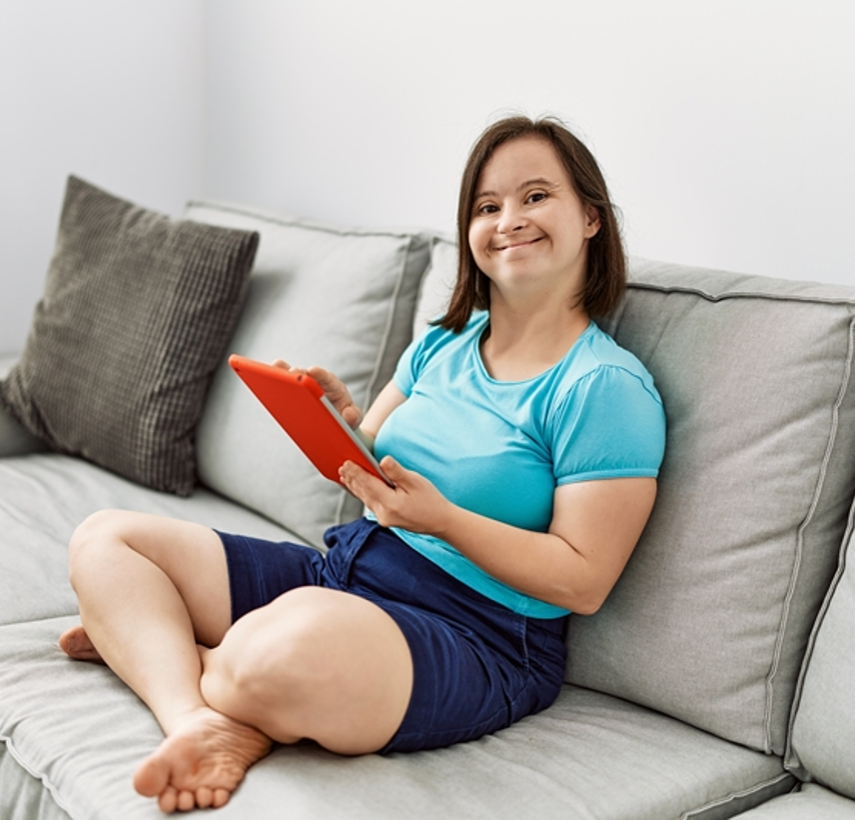 a young adult woman wearing shorts and a light blue blouse sits on a gray sofa, holding an iPad in a red case.