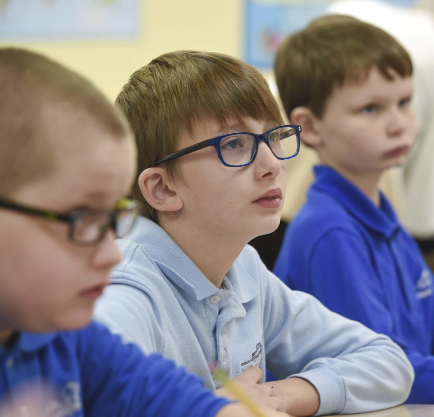 Three elementary school aged boys sit side by side in a classroom, looking forward attentively. The boys on the left and in the center are wearing glasses.