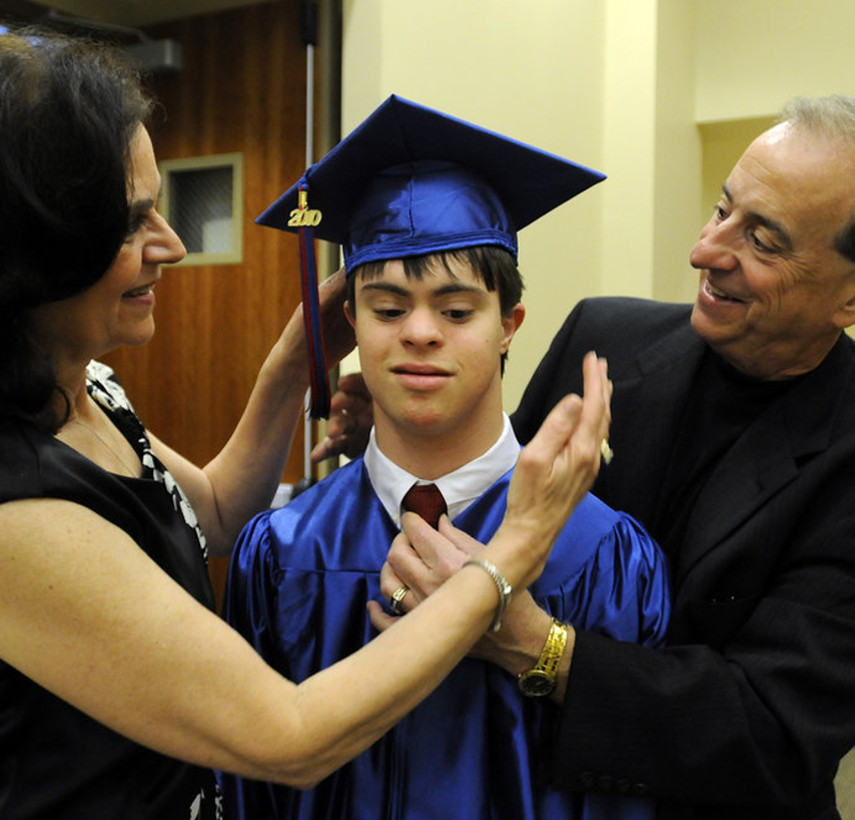 a male teenager wears a blue graduation cap and gown, while his mom on the left and his dad on the right fuss over his cap and tie.
