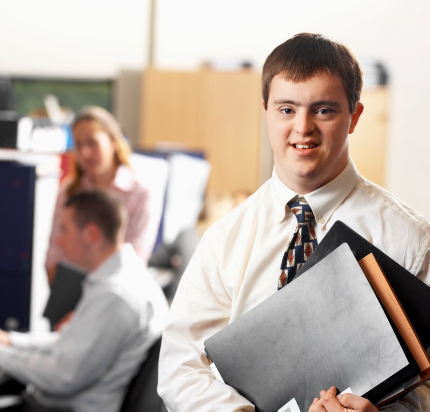 a young adult man with Down syndrome is shown in an office setting. He is wearing a white dress shirt, an tie, and holds some folders in his hands.