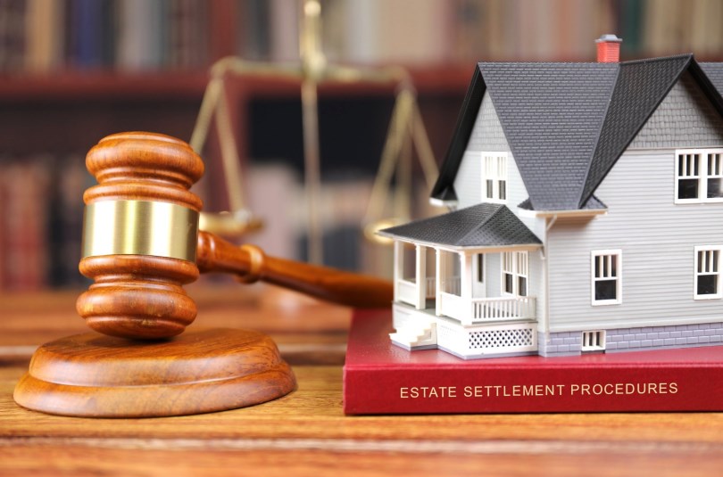 phto of a gavel and scales of justice on a wooden table, next to a model of a house sitting on a book with the title Estate Settlement Procedures