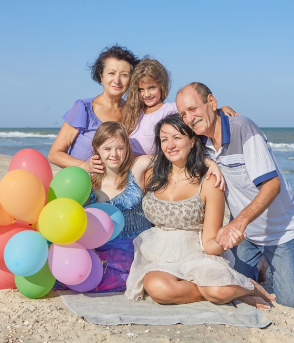 Family photo of grandma and grandpa, mom, and two daughters posed together on a blanket on the beach. A bouquet of balloons sits next to the family.