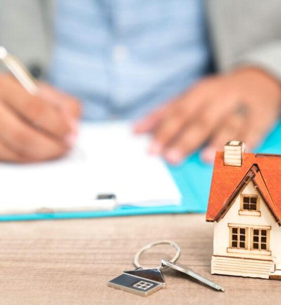 photo-in the foreground a key on a keychain and a model of a house sit on a wood desktop. In the background there is a blurred image of a man sitting at the desk, his hands are on some paperwork and he holds a pen in his right hand, signing a document.