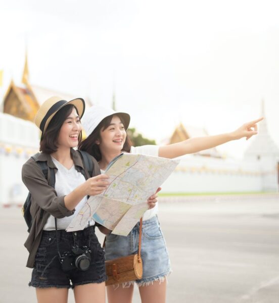 two Asian women are shown smiling on some sort of a trip. One woman holds a map, while the other points off to the right.