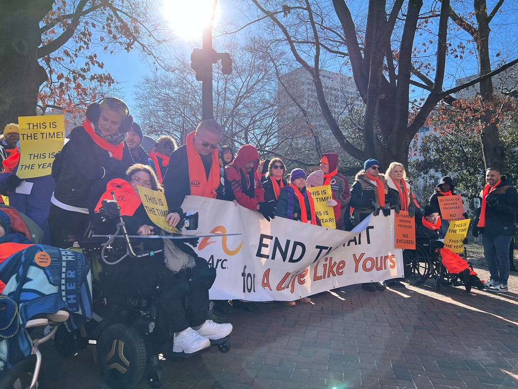 a crowd of IDD advocates in winter gear gather on the grounds of the Virginia state capitol, behind a large banner that says, 