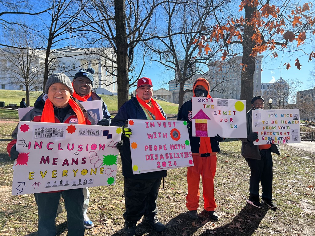 Four advocates are shown outdoors on the grounds of the Virginia state capitol, wearing winter gear and holding signs to influence legislators to support measures that benefit people with disabilities.