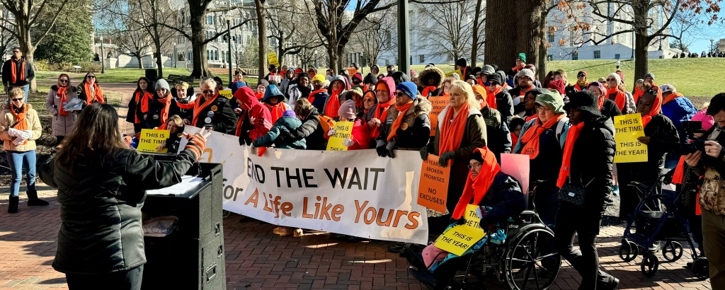 a speaker stands at a lecturn, addressing a crowd of IDD advocates in winter gear gathered on the grounds of the Virginia state capitol, behind a large banner that says, 