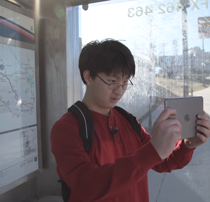a young adult asian male stands inside a bus shelter, looking at his iPad, which he holds in front of him with both hands.