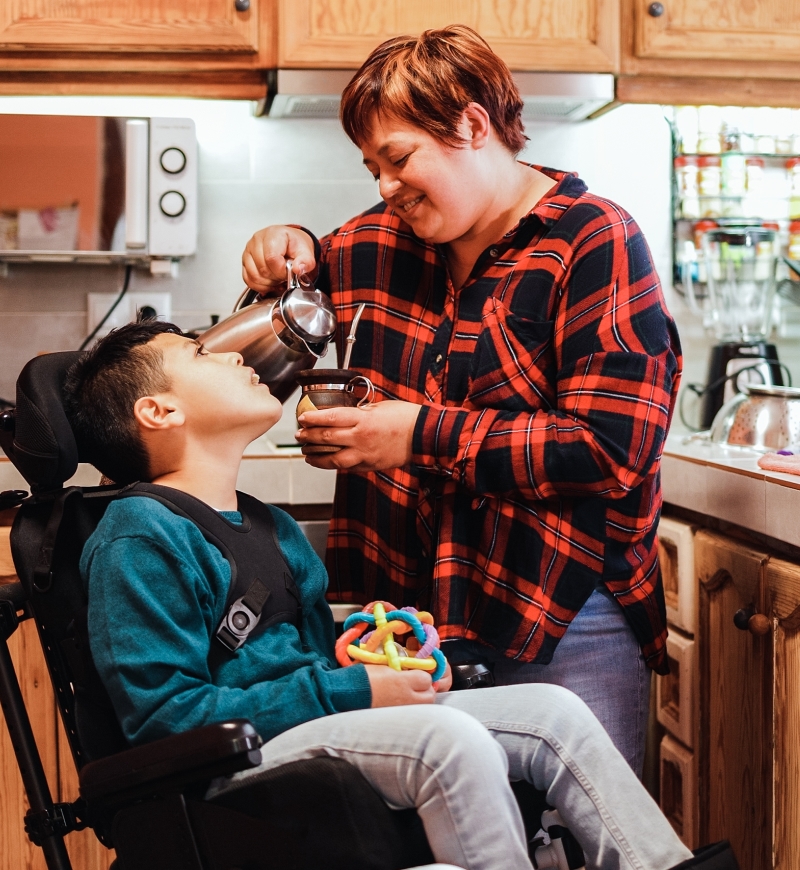 photograph of a mother and her disabled son in the kitchen. Mom is wearing a red and black checkered flannel shirt, and pours a cup of coffee, Her son is in a wheel chair, wears a teal sweatshirt, and is looking up at his mom