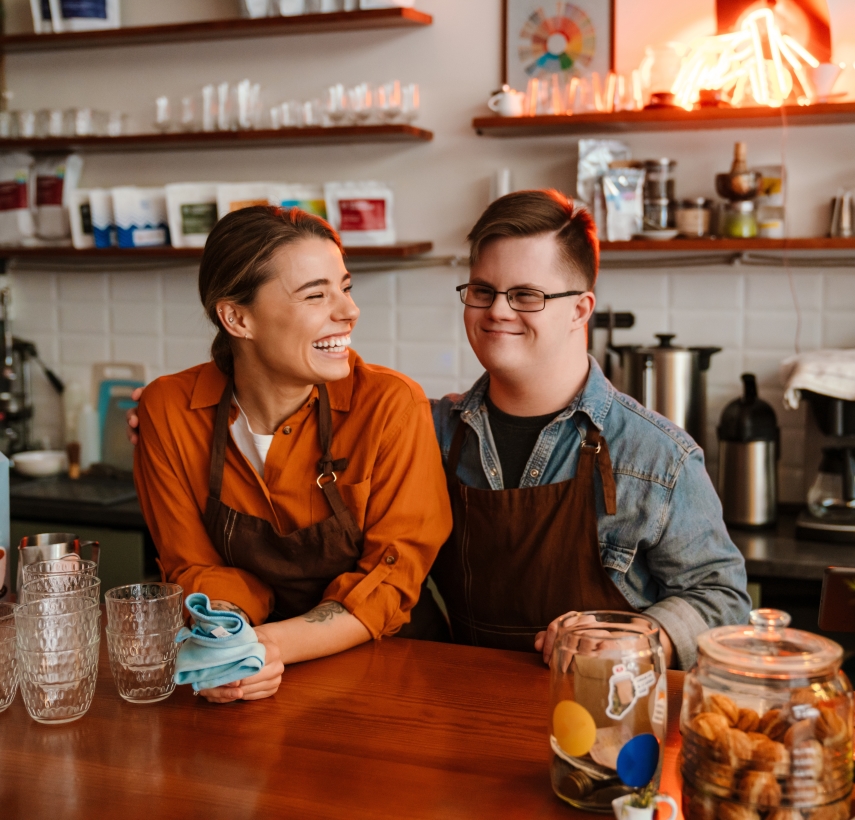 a young adult male with down syndrome and his female supervisor, both wearing brown aprons, are standing and smiling behind the counter of a coffee shop.