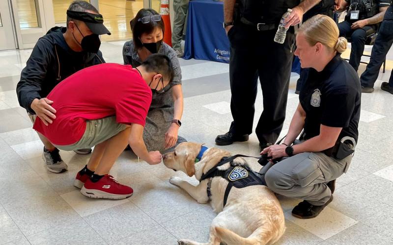 a young man kneels to pet a police dog, lying on the floor. The dog's handler and the man's parents are also shown in the photo