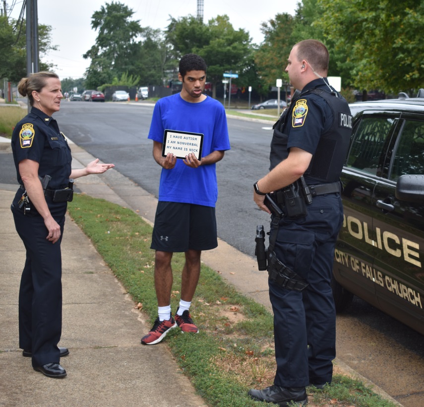 two police officers, stanind on the sidewalk next to the police car speak with a young man with autism, showing the officers an ipad screen