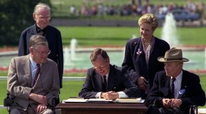 President George H. W. Bush sits at a table on the south lawn of the White House, signing a bill, with 4 onlookers, two standing, two sitting in wheelchairs.